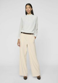 Blusa bianca a maniche lunghe con motivo a zigzag e scollatura arricciata, abbinata a pantaloni a gamba larga crema a vita alta e cintura marrone.