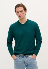 Teal gebreide sweater met een V-hals, lange mouwen en ribgebreide boorden, gecombineerd met lichtblauwe spijkerbroek. Minimale textuur en geen zichtbare patronen.