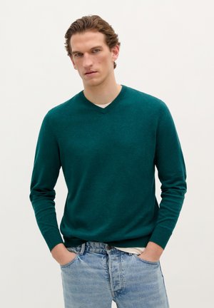 Teal gebreide sweater met een V-hals, lange mouwen en ribgebreide boorden, gecombineerd met lichtblauwe spijkerbroek. Minimale textuur en geen zichtbare patronen.