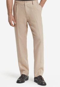 Beige bukser med en glat tekstur, der har en flad front, to sidelommer og en skræddersyet pasform. Båret med sorte lædersko.