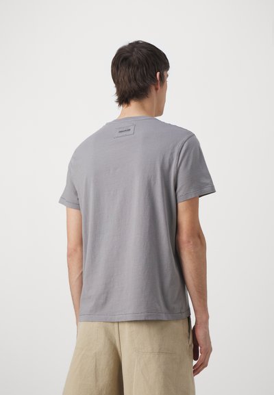 T-shirt in cotone grigio con maniche corte e collo a giro. Presenta un'etichetta rettangolare sul retro vicino al collo. Texture liscia.