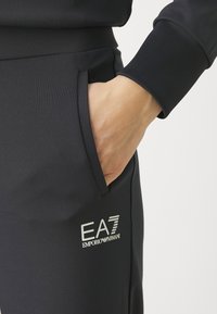 Tmavomodré tepláky s hladkým, elastickým materiálom, bočnou vreckom a reflexným logom EA7 Emporio Armani na stehne.