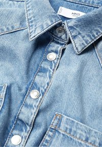 Chemise en jean bleu clair avec col pointu, boutons-pression argentés et coutures visibles. Deux poches avant incluses.