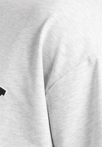 Tela de algodón gris claro con una textura suave y un estiramiento visible. Presenta un detalle de costura diagonal y un acento con el logo negro.