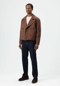 Bruine dubbelgeknoopte jas met brede revers, zijzakken en een losse pasvorm. Draagt over een lichte trui en gecombineerd met donkere spijkerbroek.