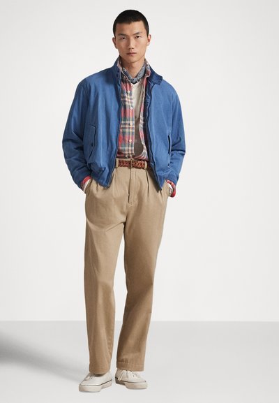 Μπλε ελαφρύ μπουφάν πάνω από πουκάμισο με καρό σχέδιο, σε συνδυασμό με μπεζ παντελόνι και λευκά αθλητικά παπούτσια, με πλεκτή ζώνη και φουλάρι με σχέδιο στο λαιμό.