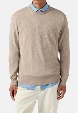 Uomo che indossa un maglione beige a girocollo sopra una camicia azzurra con bottoni e pantaloni color crema, in piedi davanti a uno sfondo semplice.