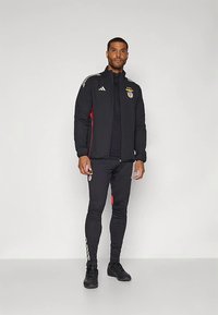 Musta urheilutakki, jossa on punaisia yksityiskohtia ja kultaiset raidat, logolla varustettuna. Yhdistetty mustiin slim-fit-housuihin ja mustiin kenkiin.