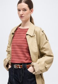 Γυναίκα με ανοιχτόχρωμο δέρμα και καστανά μαλλιά σε αλογοουρά, ντυμένη με μπεζ καμπαρντίνα, κόκκινη ριγέ μπλούζα, σκούρα τζιν ψηλόμεσα και καφέ ζώνη με τα χέρια στις τσέπες.