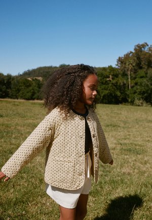 Fată tânără cu păr creț, purtând o jachetă bej cu buline și pantaloni scurți albi, stă cu brațele întinse într-un câmp însorit cu iarbă.