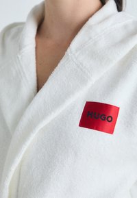 Balta vonia su minkšta tekstūra, turinti raudoną stačiakampį pleistrą su juodu užrašu "HUGO" ant apykaklės.