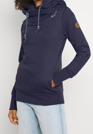 Osoba ubrana w granatową bluzę z kapturem z kołnierzem kominem i sznurkami, ręce w kieszeniach, w połączeniu z jasnoniebieskimi dżinsami.