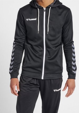 Homme portant une veste de sport Hummel noire avec une fermeture éclair blanche et des motifs en chevrons sur les manches, assortie à un pantalon noir.