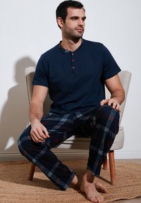 Cămașă scurtă din bumbac, de culoare albastru închis, cu guler în carouri, asortată cu pantaloni de pijama în carouri, în nuanțe de albastru, roșu și alb. Se află așezat pe un scaun.