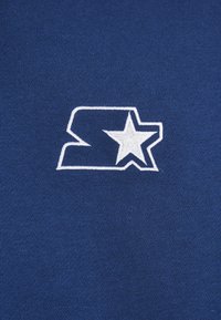 Tessuto blu navy con maglia testurizzata; presenta un logo ricamato bianco che rappresenta una "S" intrecciata con una stella.