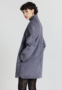 Donna con i capelli ricci e corti, che indossa un cappotto grigio oversize e leggings neri in pizzo, in piedi di lato con le mani nelle tasche su uno sfondo semplice.