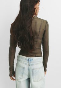 Gennemsigtig langærmet top med et tekstureret olivengrønt design, parret med lyseblå højtaljede jeans med baglommer.