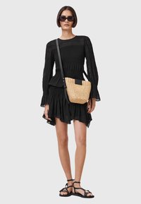 Schwarzes Smock-Kleid mit langen Ärmeln, geschichteter Säume und Rüschendetails. Geflochtene Strohhüfttasche mit einem schwarzen Träger. Schwarze Sandalen mit Riemen.