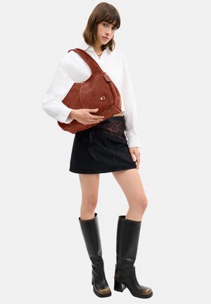 Vrouw draagt witte blouse, zwarte rok, hoge zwarte laarzen, houdt grote bruine suède schoudertas tegen een effen achtergrond.