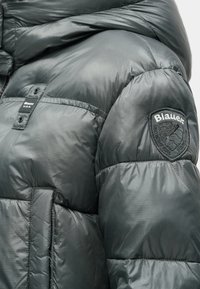 Σκούρο γκρι μπουφάν με φούσκωμα, που διαθέτει γυαλιστερή, ραμμένη υφή, φερμουάρ και κυκλικό λογότυπο στο αριστερό ώμο.
