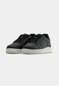 Juoda pora žemų sportbačių su baltais padais ir juodomis raištelėmis, sudėliota šalia ant paprasto šviesaus fono.