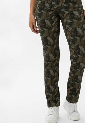 Personne portant un pantalon à motif camouflage et des baskets blanches, debout devant un fond blanc uni.