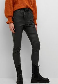 Svarta läderlook-byxor med elastisk midja och knytstängning, figurnära passform, i kombination med svarta ankelboots och en orange tröja.
