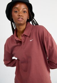 Bordeaux dlouhý rukáv polo tričko s knoflíkovou légou a logem Nike. Model nosí černý klobouk typu bucket. Hladká textura látky.
