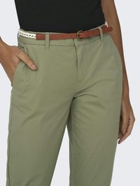 Gros plan sur une personne portant un pantalon vert clair avec poches latérales, un haut noir, et une ceinture fine marron avec une boucle ronde dorée.