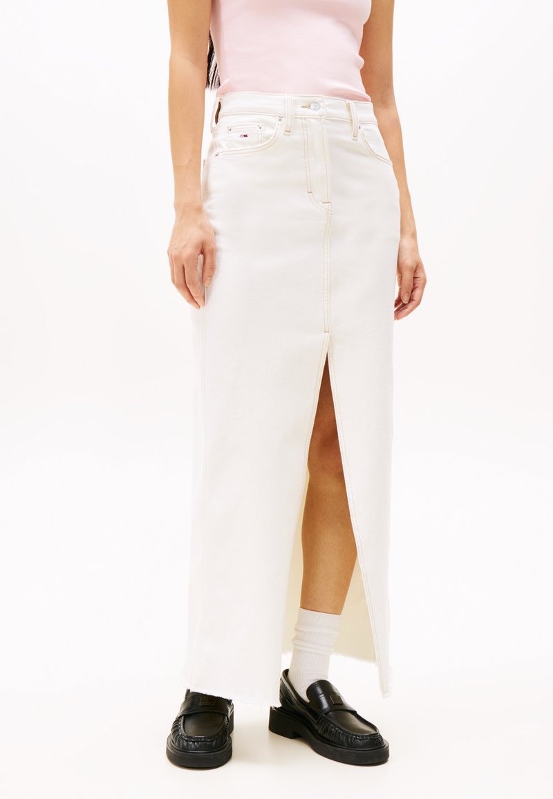Jupe maxi en denim blanc avec une fente sur le devant, dotée d'une taille haute, de coutures contrastées et d'un ourlet brut. Associée à des chaussures noires et des chaussettes blanches.