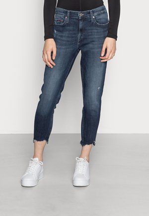 Person trägt dunkelblaue, verkürzte Jeans mit ausgefranstem Saum und leichter Used-Optik sowie weiße Plateausneaker und steht vor einem schlichten Hintergrund.