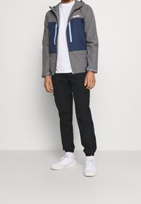 Herrgrå jacka med blå detaljer, dragkedjefickor och huva, kombinerad med svarta cargobyxor och vita sneakers. Mjuk tygtextur.