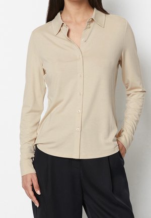 Kobieta ubrana w beżową koszulę na guziki z długim rękawem, włożoną do czarnych spodni, stoi z jedną ręką w kieszeni na białym tle.