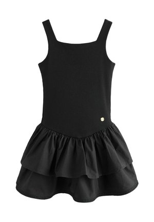 Niña joven con cabello rubio largo vistiendo un vestido negro sin mangas con falda de capas y zapatos Mary Jane dorados, de pie sobre un suelo gris.