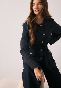 Tumši zila jaka no strukturētas auduma, ar pieguļošu dizainu, garām piedurknēm, lielām dekoratīvām pogām un aizdari viduklī.