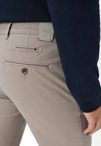 Beige cotton trousers featuring two back pockets with horizontal openings, a branded label in gray, and a button closure.