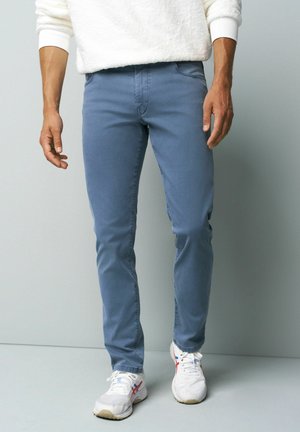 Man draagt een slim fit blauwe broek, witte sneakers met rode en blauwe accenten, en een wit getextureerd shirt met lange mouwen tegen een effen grijze achtergrond.