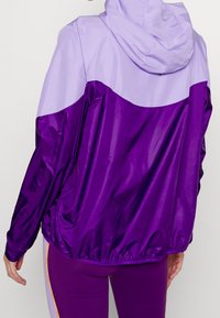 Veste à capuche violette et lavande au fini brillant, avec un ourlet serré et des blocs de couleur contrastante. Matériau léger.