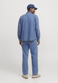 Camisa de mezclilla y pantalones a juego en azul, con detalles de botones y bolsillos. El conjunto se complementa con una gorra marina y calzado beige.