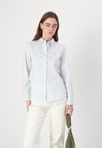 Camicia a righe blu chiaro con maniche lunghe, colletto e piccolo dettaglio del logo, abbinata a pantaloni bianchi e una borsa verde.