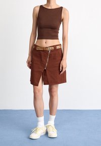 Top corto marrone, gonna di jeans marrone con cerniera e cintura decorata, abbinata a calzini bianchi e sneakers gialle con accenti blu.