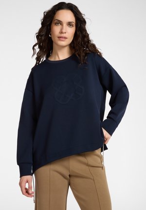 Donna con capelli ricci e scuri che indossa una felpa oversize blu navy con un motivo in rilievo e pantaloni beige, in piedi su sfondo bianco.