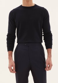 Marineblauer Kaschmirpullover mit geripptem Rundhalsausschnitt und enganliegenden langen Ärmeln, kombiniert mit maßgeschneiderten, dunklen Hosen, die ein Ein-Knopf-Detail aufweisen.