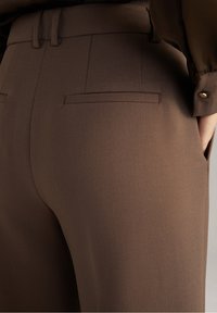 Gros plan sur un pantalon marron sur mesure avec des poches passepoilées à l'arrière et la main d'une personne dans la poche latérale, portant un chemisier à manches longues avec un poignet boutonné.