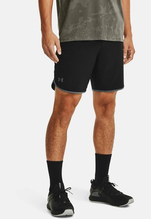 Homme portant un short de sport noir avec un liseré gris, des chaussettes noires montantes et des chaussures de course noires, debout les mains détendues le long du corps.