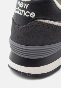 Pätka čierneho a sivej športovej obuvi New Balance so semišovou textúrou, rebrovanou medzipodrážkou a bielym logom značky na golieri päty.