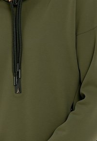 Tessuto di felpa verde oliva con una texture liscia, caratterizzato da un laccio nero con puntali metallici e una tasca con zip sul lato.