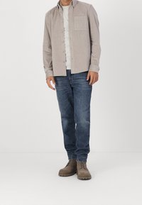 Camisa de boton de pana beige con bolsillo de parche, camiseta blanca debajo, combinada con jeans de mezclilla azul y botas de cordones marrones.