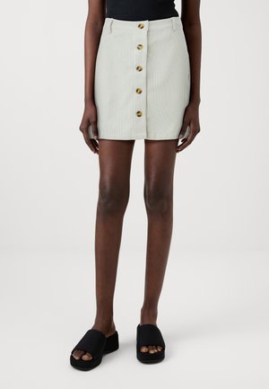 Kobieta w jasnobeżowej mini spódnicy zapinanej z przodu na guziki, w czarnych sandałach na koturnie i czarnej bluzce bez rękawów.