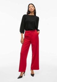 Top noir en tricot avec un motif chevron texturé, associé à un pantalon rouge à jambes larges et des escarpins noirs à bout pointu. Design droit dans l'ensemble.
